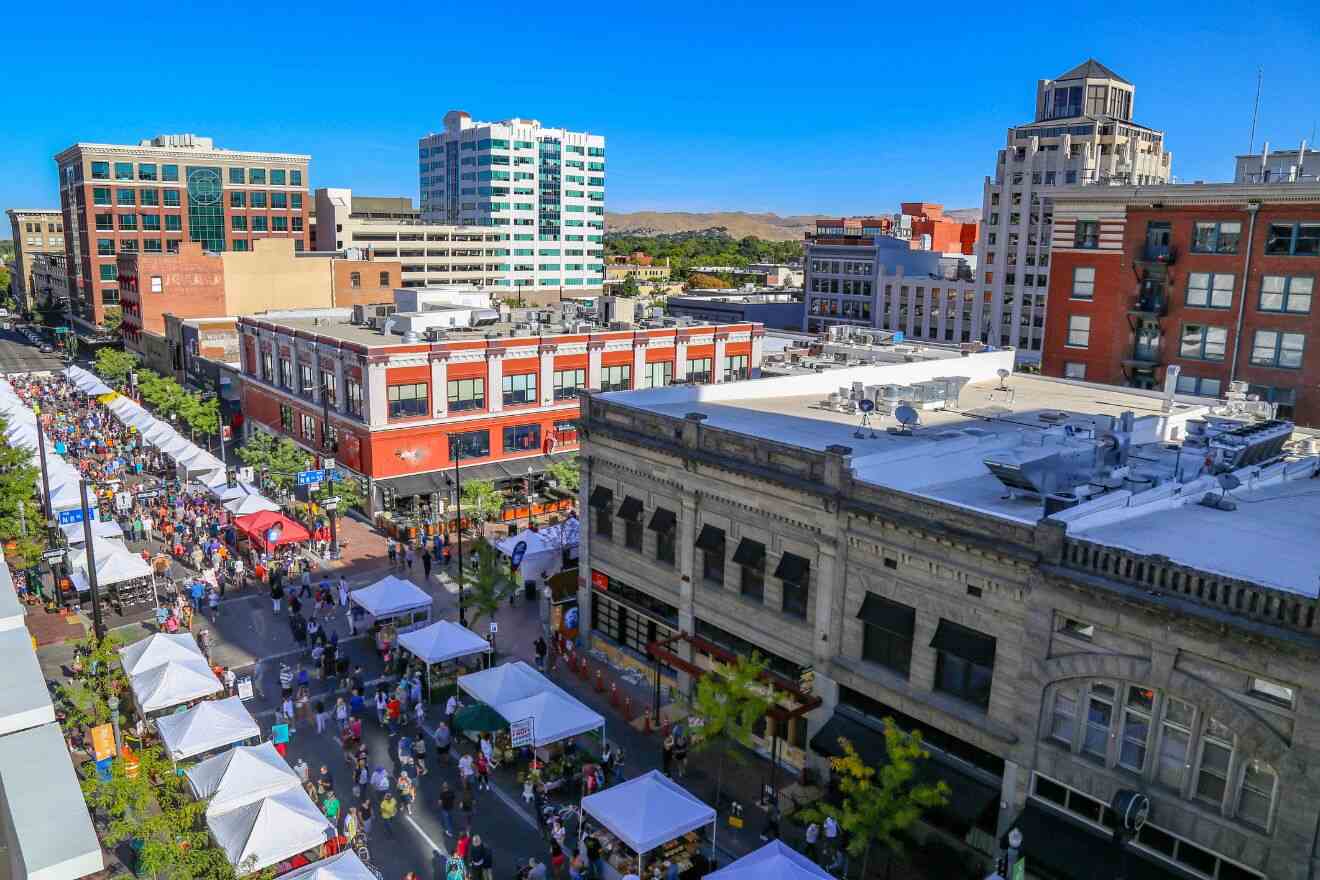 Aerial view of a bustling outdoor market in a city, with numerous white tents lining the street, buildings of various styles, and people walking and shopping under a clear blue sky.