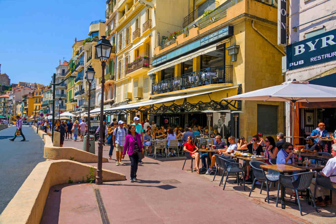 A lively street scene with people dining at outdoor cafes and walking along the sidewalk in front of colorful buildings under a clear blue sky.