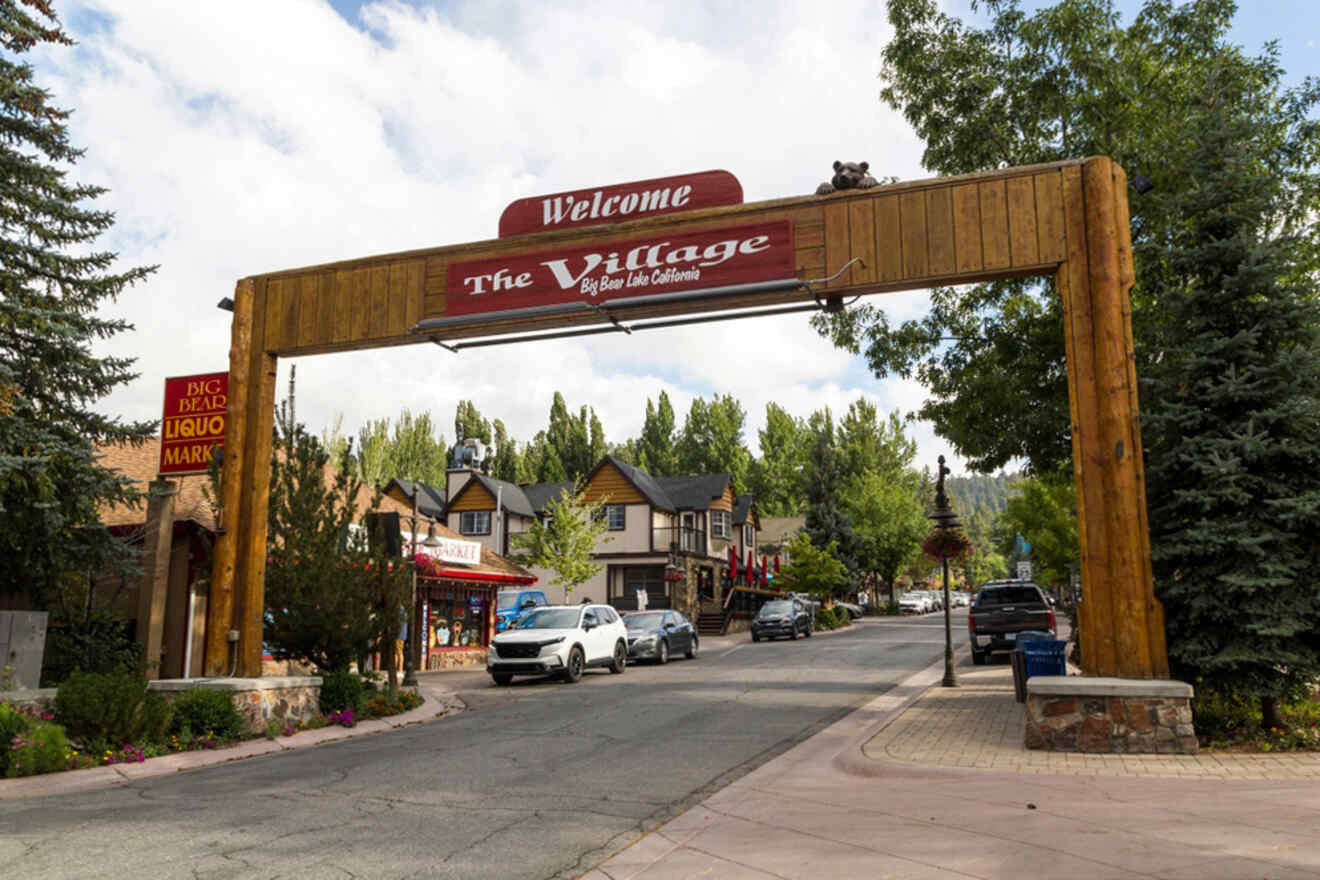 An entrance archway labeled "The Village" seen in a small town street lined with shops, trees, and parked cars.