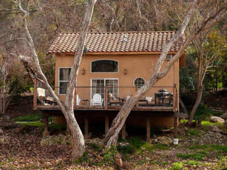 A small tan house with a red tiled roof, elevated on stilts, is surrounded by trees. There is a furnished wooden deck in front.