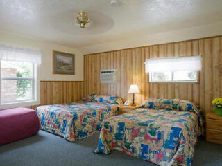 A modest motel room with wood-paneled walls, two beds covered in vibrant, patterned bedspreads, a nightstand with a lamp, two windows, an air conditioner, a framed picture, and a maroon bench.