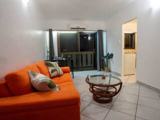 A living room with a bright orange couch, a glass coffee table, a small dining table with two chairs, and a large window with dark curtains. The room has white walls and tile flooring.