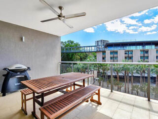 A balcony with a wooden table, benches, a ceiling fan, and a barbecue grill, overlooking a view of buildings and trees.