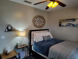 A well-kept bedroom with a bed featuring blue and gray bedding, a nightstand with a lamp, decorative wall art, a ceiling fan with light, and a small shelf with decor items.