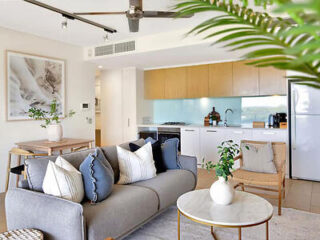 Modern living room with a gray sofa, blue and white cushions, wooden furniture, and a potted plant. The open kitchen in the background features light wood cabinets, an island, and stainless appliances.