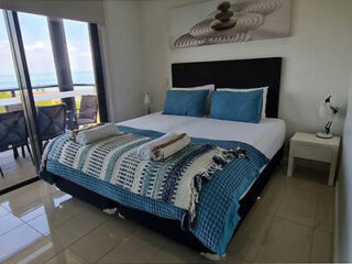 A neatly made bed with blue and white bedding, flanked by two matching nightstands and lamps. A sliding glass door leads to a balcony with a view of the ocean. A stone arrangement artwork hangs above the bed.