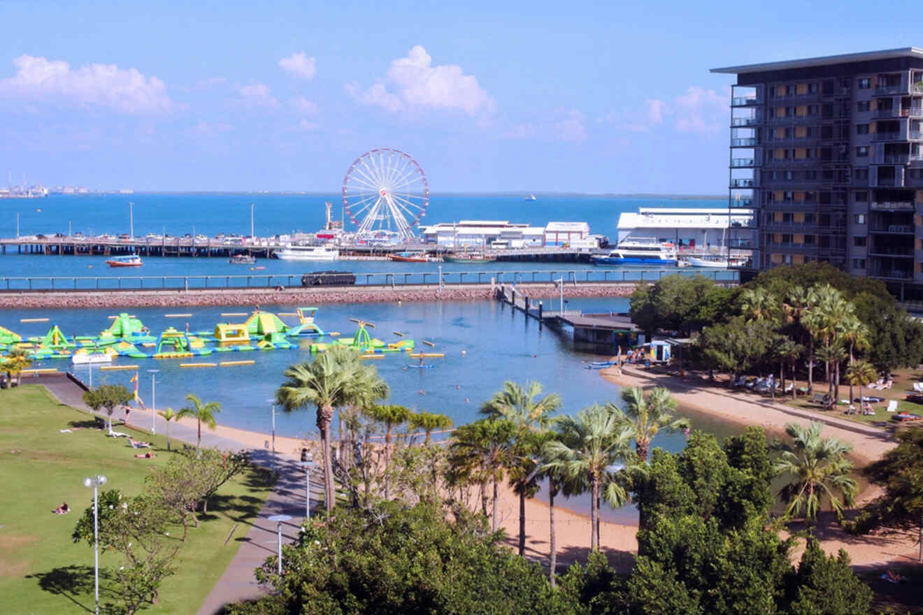 A coastal scene featuring a Ferris wheel, waterfront buildings, inflatable water park, and palm trees. Green spaces and a sandy beach are in the foreground, with a calm blue sea in the background.