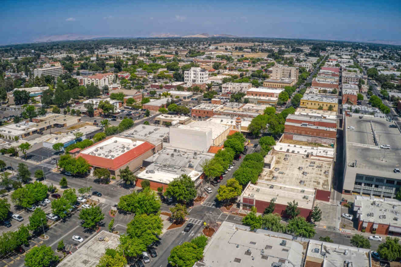 Aerial view of a city with a mix of low-rise buildings, tree-lined streets, and a parking lot. The landscape is expansive under a clear blue sky.