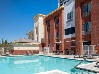 Outdoor swimming pool area with lounge chairs and umbrellas, adjacent to a brick and stucco building under a clear blue sky.