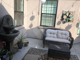 A small outdoor patio with a cushioned loveseat, a wicker table, a water fountain, a hanging plant, string lights, and a decorative rug.