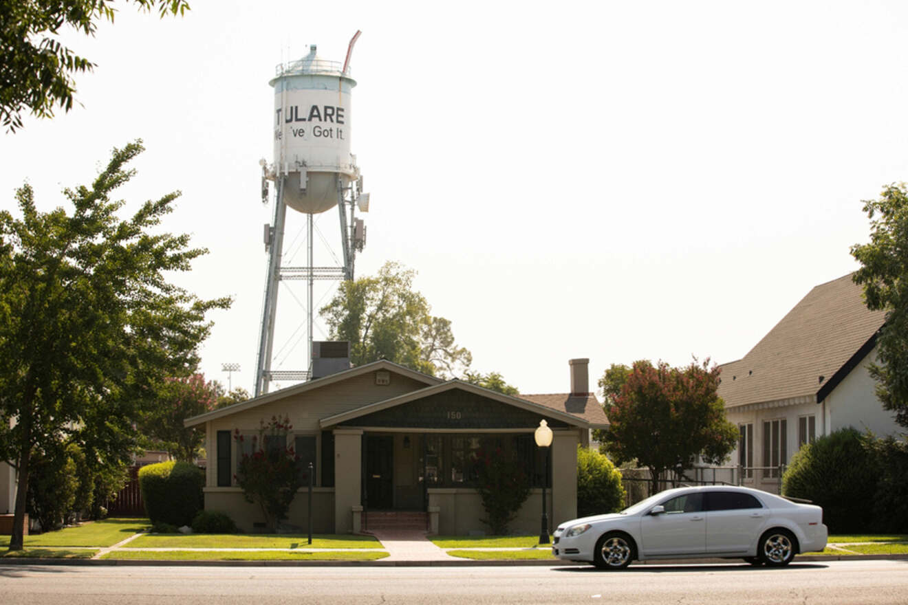 A small house with a porch sits next to a white car on a tree-lined street. A tall water tower in the background displays the words "Tulare - We've Got It.