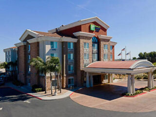 Exterior view of a four-story hotel with a covered entrance, arching design, and visible signage. The hotel features brick and stucco construction, palm trees, and a clear blue sky in the background.