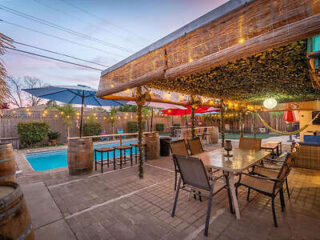 Outdoor patio area with a dining table, chairs, and string lights. Nearby, there are umbrellas, a bar area, and a swimming pool surrounded by lounge chairs. The sky is dusk.