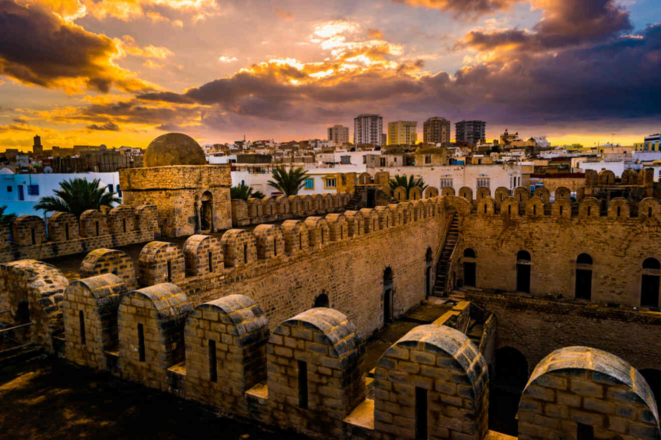 A dramatic sunset over the ancient stone walls of a fortress in Tunis, with modern buildings in the background under a vibrant sky filled with clouds