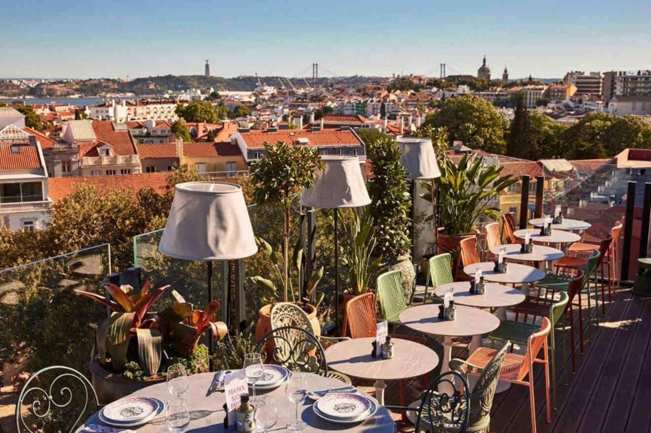 Rooftop restaurant with tables and chairs overlooks a city, featuring a variety of plants and lamps, under a clear sky.