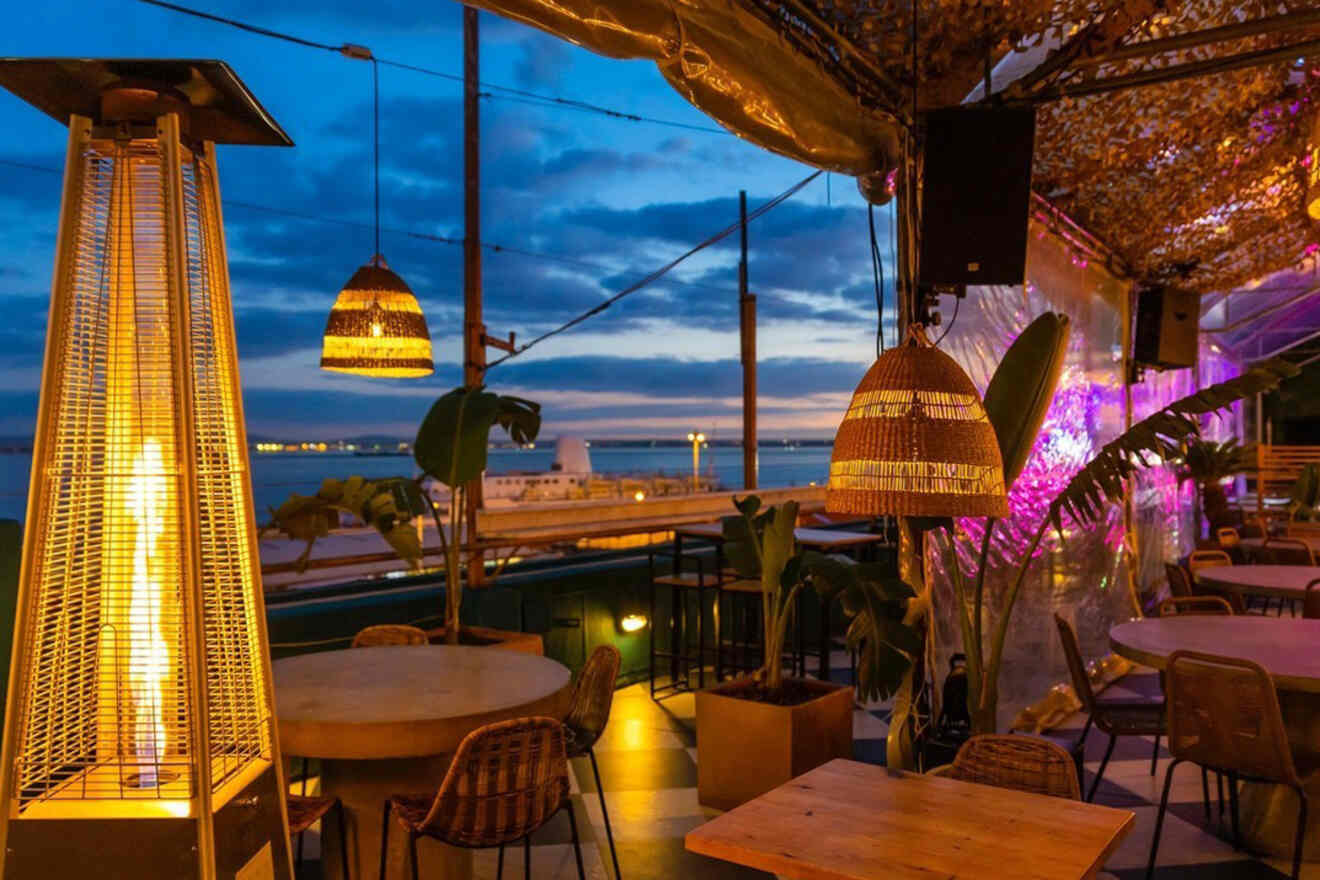 Outdoor restaurant seating area with wicker lamps and a tall heater, overlooking a view of the sea at sunset. Tables and chairs are arranged under a canopy with ambient lighting.