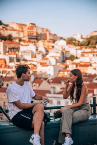 Two people sit on a wall overlooking a cityscape with colorful buildings. The person on the left wears a white T-shirt and black shorts, while the person on the right wears a beige top and green trousers.