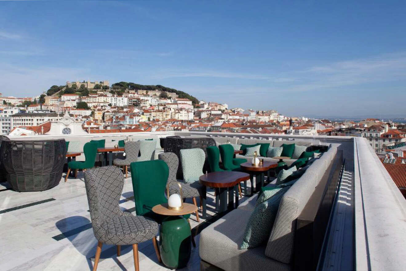 A rooftop terrace with green and gray seating overlooks a city with red-roofed buildings and a hilltop castle in the background under a clear blue sky.