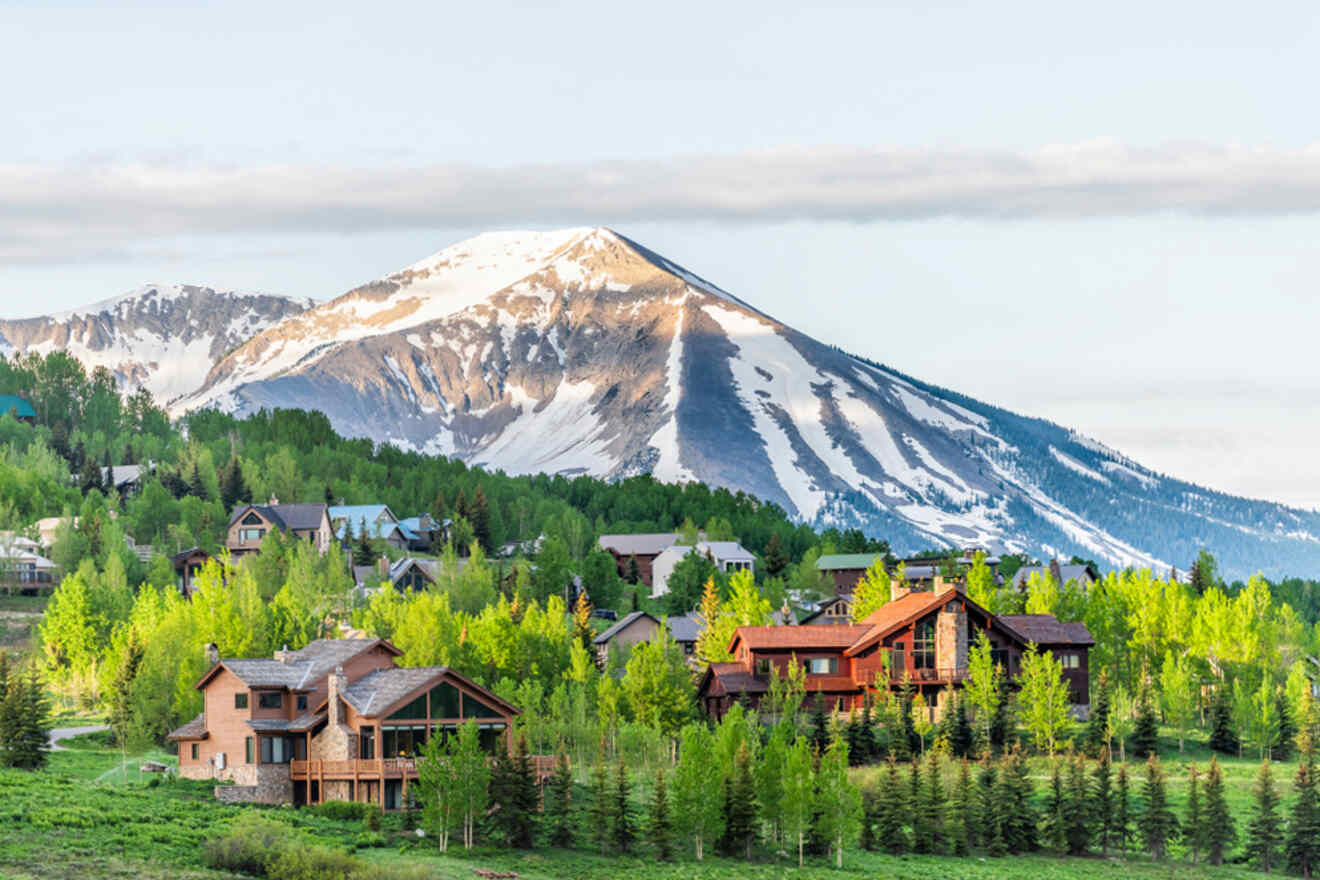 A scenic mountainous landscape with several wooden houses surrounded by green trees. Snow-capped peaks dominate the background under a partly cloudy sky.