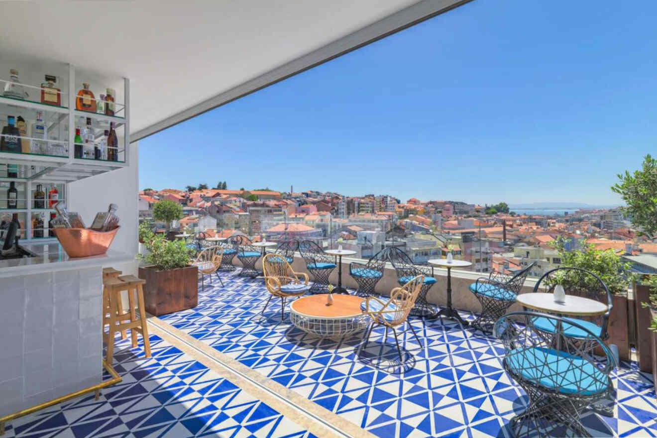 An outdoor rooftop bar with a geometric blue-and-white tile floor, wicker chairs, and small round tables overlooking a cityscape under a clear blue sky.