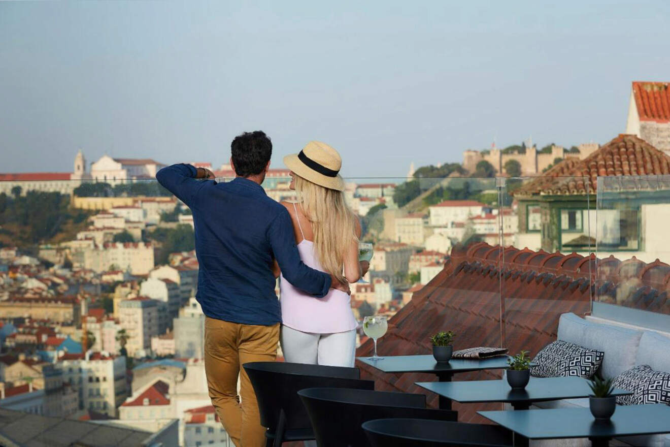 Two people stand on a rooftop terrace overlooking a cityscape, with the person on the left taking a photo.