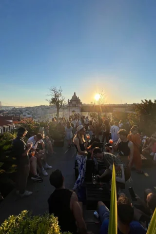 A crowd of people gathered on a rooftop terrace at sunset, enjoying drinks and socializing. The view includes a distant cityscape and a prominent domed building under a clear sky.