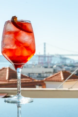 A glass of iced red cocktail with an orange slice garnish sits on a table outdoors, with a distant view of a bridge and rooftops in the background.