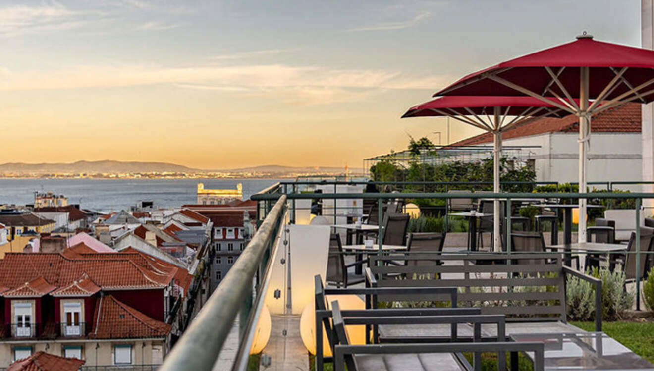 A rooftop terrace with red umbrellas, tables, and chairs overlooks a cityscape with tiled roofs and a body of water in the background at sunset.