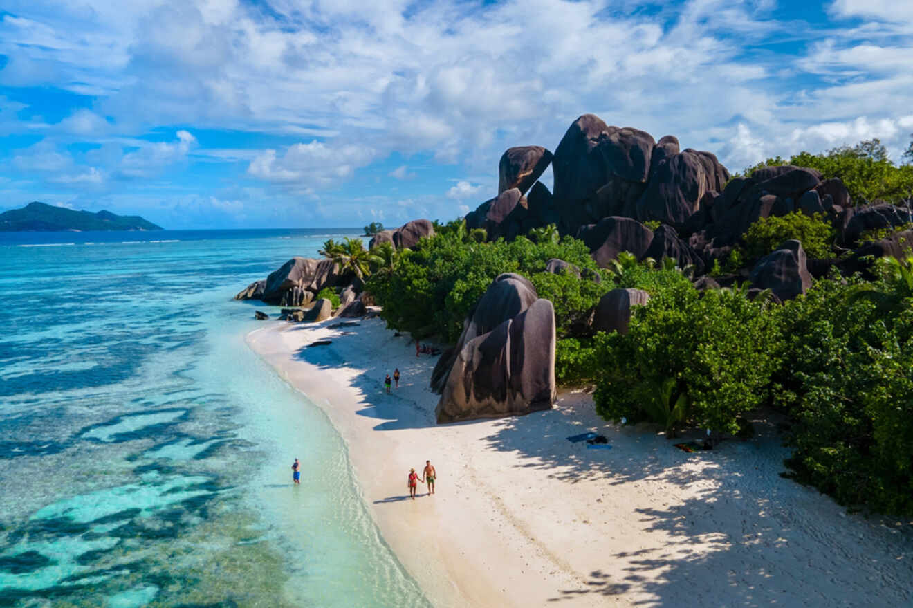 A secluded beach with white sand, large granite boulders, and lush greenery, with clear blue water stretching to the horizon.