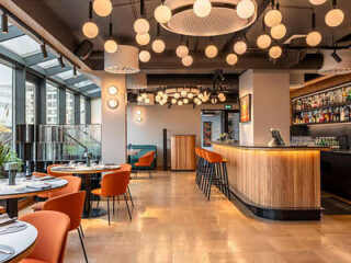 Modern restaurant interior with orange chairs, round tables, wooden bar counter, and spherical ceiling lights. Large windows line one wall.