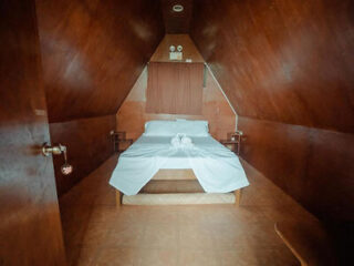 A small, wooden, A-frame cabin bedroom with a single, neatly made bed adorned with white sheets and two towel swans on top. The room has slanted walls and a brown color scheme.