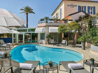 Outdoor pool area of Hotel Le Suquet in Le Cannet, featuring lounge chairs with towels, umbrellas, and a view of the building and surrounding palm trees.