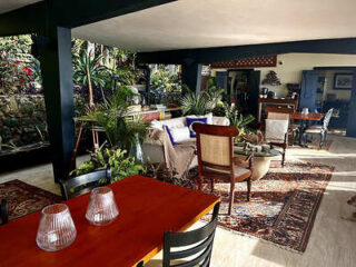 A spacious indoor seating area featuring a wooden dining table, several chairs, plants, rugs, and a couch under a covered patio, with surrounding greenery.