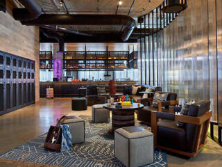 A modern lounge area with a bar, sleek seating, and lockers. The space features a mix of contemporary decor, including a patterned rug, stools, and industrial-style lighting and ceiling pipes.