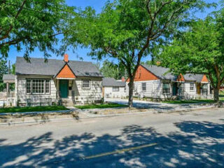 A row of small, single-story houses with gray siding and red roofs is shaded by tall green trees along a quiet street on a sunny day.