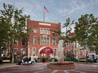 A historic brick building with an awning reading "The Redbury" sits in the background. A roundabout in the foreground features a tall stone monument and surrounding flowers. A golf cart is parked nearby.