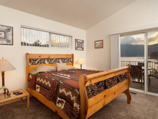 A cozy bedroom with a wooden bed, patterned bedding, bedside table with lamp, and sliding glass doors leading to a balcony with a view of a lake and mountains.