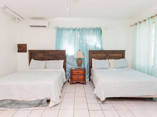 A bedroom with two double beds, matching bedding, a central nightstand with a lamp, and light blue curtains. The room has tiled floors and a wall-mounted air conditioner.