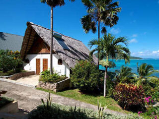 A charming cottage with a thatched roof, surrounded by tropical plants, overlooking the ocean with bright blue water.