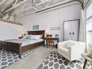 Modern bedroom with a large wooden bed, white armchair, small desk and chair, industrial ceiling, and patterned rugs on the concrete floor. Room has white walls and a window with natural light.