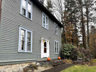 Two-story gray house with blue trim, white door, and two pumpkins on the front steps. A "For Sale" sign is visible in the yard. Trees and foliage are in the background.