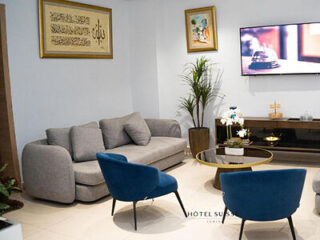 A modern living room with a grey sofa, blue chairs, and a flat-screen TV, adorned with framed Arabic calligraphy.