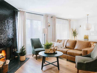 A modern living room with a brown sofa, gray armchairs, a round black coffee table, and a fireplace with a black tiled facade. Light pours in through large windows with sheer curtains.