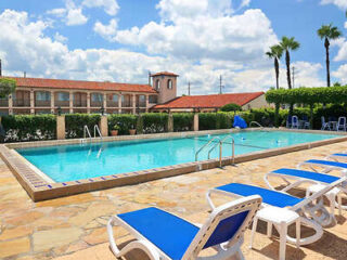 Outdoor swimming pool with blue lounge chairs on stone decking surrounding it. A building with terracotta-colored roofs and a green hedge are in the background.