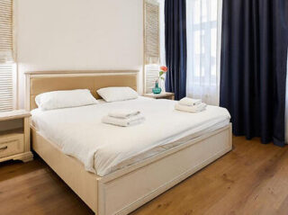 A neatly made bed with white linens and folded towels, flanked by bedside tables, in a bright room with wooden flooring and blue curtains.