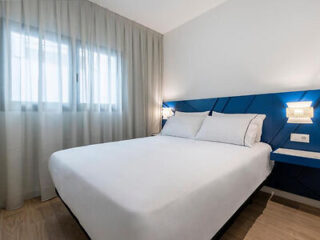 A minimalistic bedroom with a large bed, white linens, and blue accent headboard. Curtains cover a window on the left wall, and wall-mounted lamps provide lighting on either side of the bed.