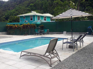 A small private pool area with lounge chairs and an umbrella, surrounded by greenery and featuring a turquoise house in the background.