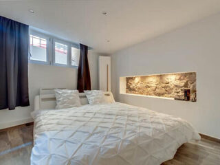 A modern bedroom with a large bed, a stone accent wall with built-in lighting, wood floors, and a small window with dark curtains.