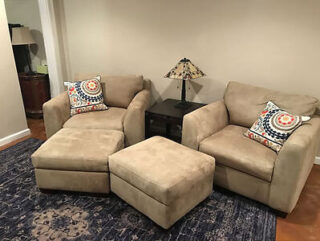 A living room setup with two beige armchairs, a matching ottoman, a small black side table between them, and a lamp on the table. The armchairs have patterned throw pillows. The floor has a dark blue rug.