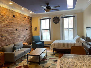 A studio apartment with a bed, sofa, armchair, coffee table, and TV. Features exposed brick wall, large windows, ceiling fan, and overhead lighting.